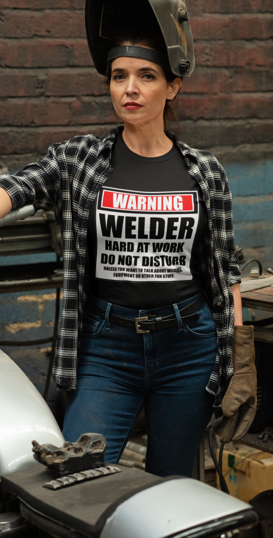Woman welder wearing a black Warning Welder Hard At Work shirt near a motorcycle.